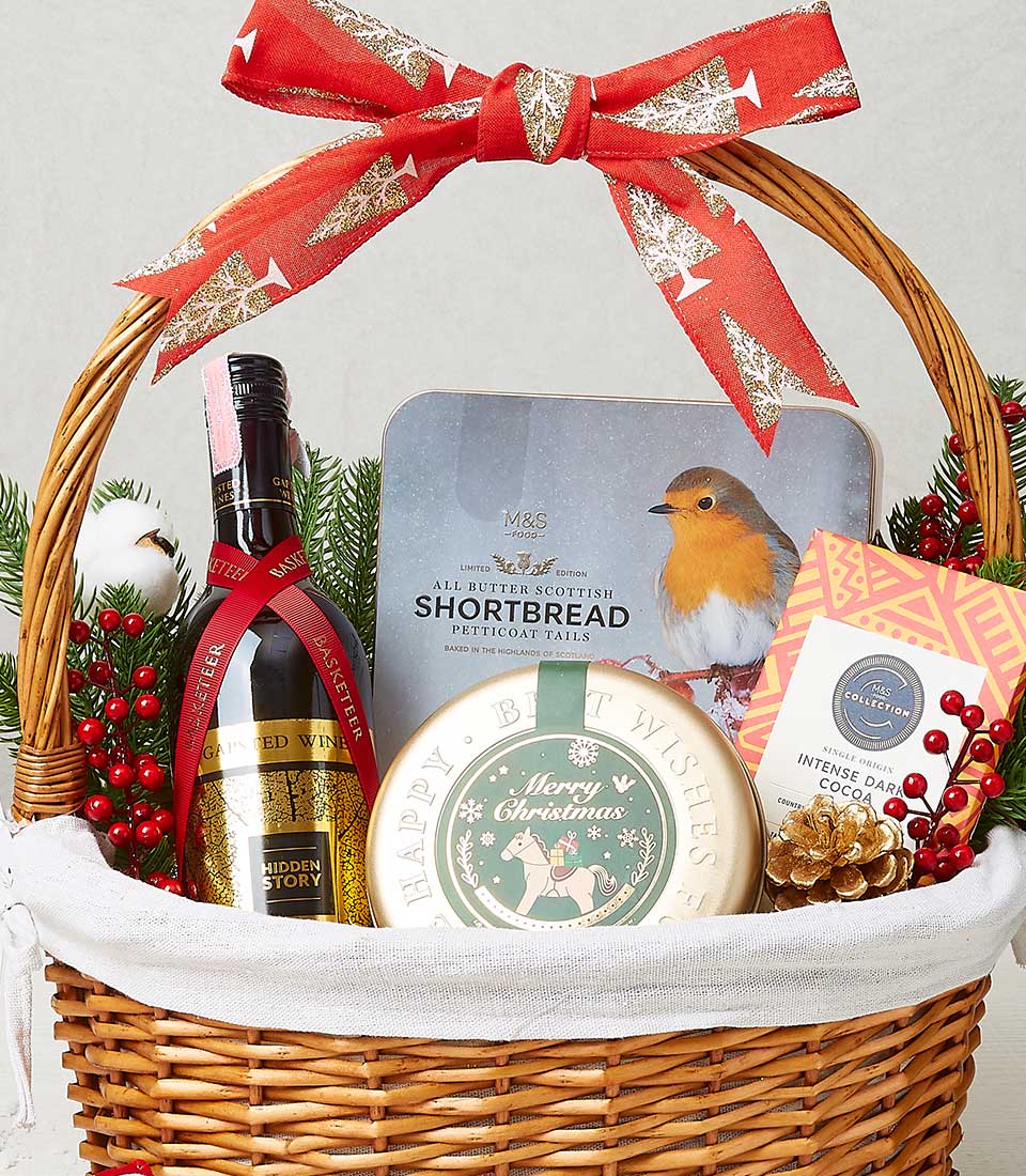An opulent wicker gift basket, elegantly adorned with a luxurious red bow, features a superb selection from Basketeer. Inside this sophisticated collection, you'll discover a distinguished bottle of fine red wine, alongside an enchanting tin of shortbread cookies graced with a festive robin design. Adding to the yuletide charm is a circular 
