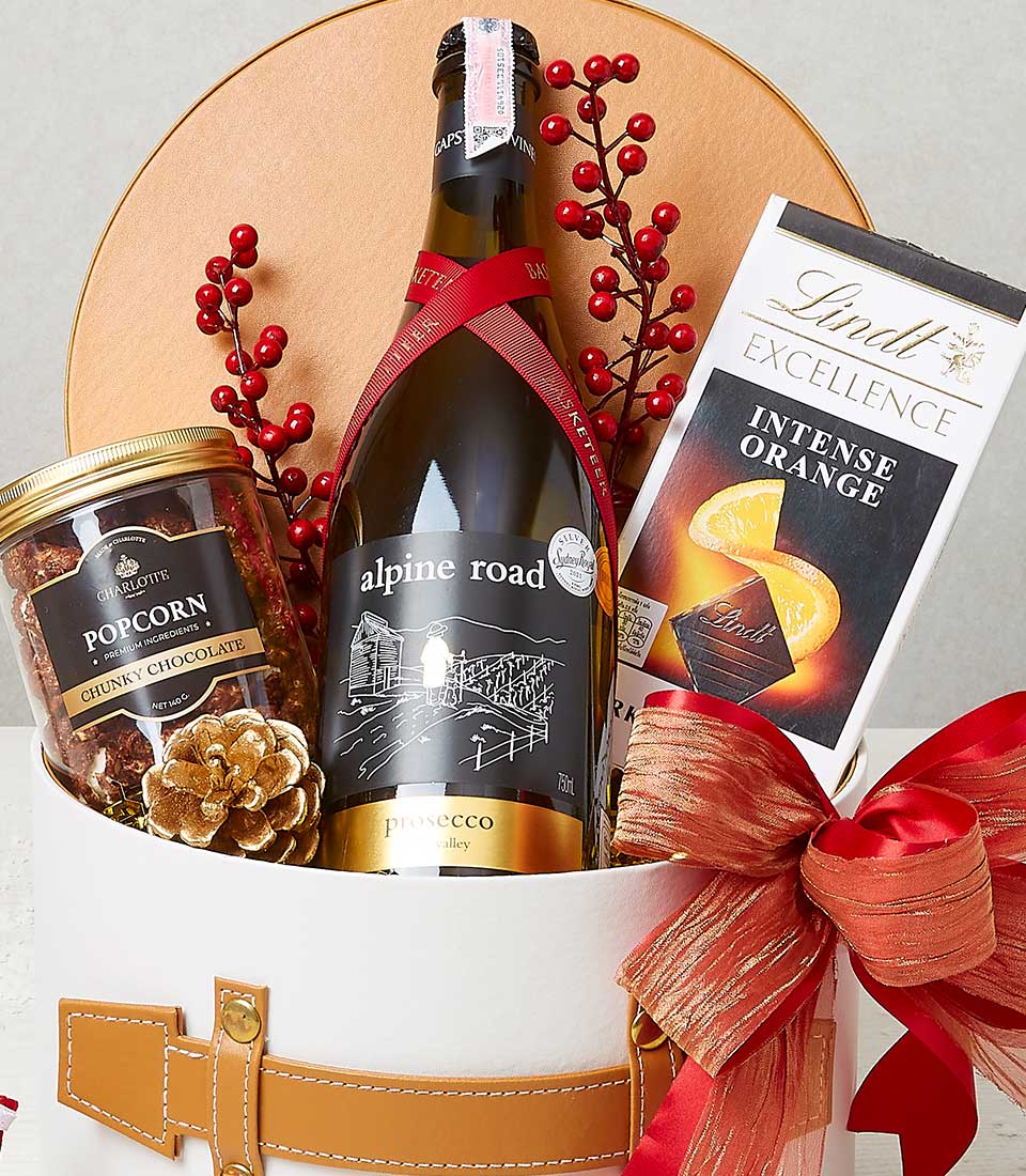 A lavish gift basket features a bottle of exquisite Alpine Road Prosecco, an indulgent Lindt Excellence Intense Orange chocolate bar, and a stylish container filled with decadent chunky chocolate popcorn. The elegant white basket is adorned with a sumptuous red ribbon, delicate festive red berry accents, and proudly showcases the Basketeer logo, embodying the spirit of luxury and celebration.