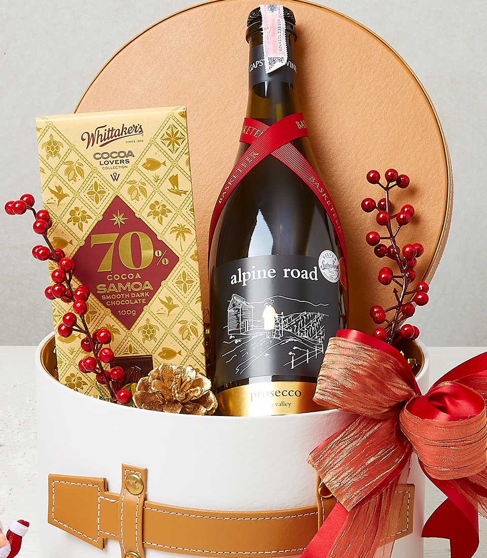 A luxurious holiday gift box exudes opulence with its bottle of Alpine Road Prosecco elegantly wrapped in a sumptuous red ribbon. Nestled beside it is a 100g Whittaker's Cocoa Lovers 70% Cocoa Samoa chocolate bar, promising rich indulgence. Accentuating the scene are festive red berries and an exquisitely gilded pinecone, serenading the spirit of the season. The box itself is a masterpiece from Basketeer, adorned with an impeccable red ribbon and topped with a delicate bow, embodying sophistication and festive charm.