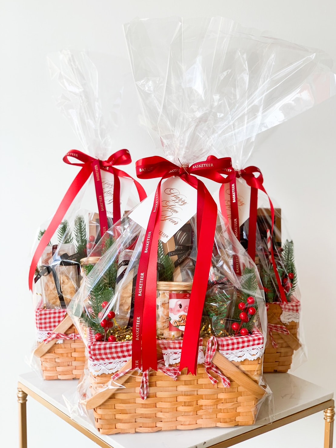 Three festive gift baskets wrapped in clear cellophane with red bows. Each basket contains holiday-themed items, including greenery, red berries, and various goodies. The baskets are placed on a white surface with a light background.