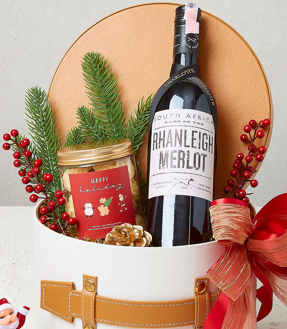 A sumptuous gift basket by Basketeer, featuring a distinguished bottle of South African Rhanleigh Merlot, elegantly nestled beside a jar of holiday jam adorned with charming illustrations of Santa Claus, reindeer, and snowman. Enhanced with lush pine branches and vibrant red berry decorations, the scene is completed by a luxurious red ribbon and an exquisite tan leather handle. The presence of the Basketeer brand logo on the ribbon adds an extra touch of opulence to this inviting festive offering.