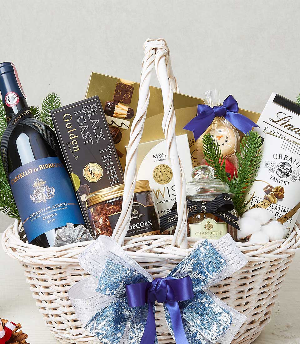 A tasteful white wicker gift basket exudes opulence, brimming with an exquisite selection of luxury items. Nestled within are a fine bottle of red wine, a decadent chocolate bar, a jar of gourmet mixed nuts, an artisanal box of biscuits, a jar of sumptuous popcorn, and indulgent chocolates. To heighten the festive charm, an elegant Christmas decoration is included. This sophisticated ensemble is adorned with a lush blue and white bow featuring the prestigious Basketeer logo prominently displayed on the ribbon, signifying impeccable taste and thoughtfulness.