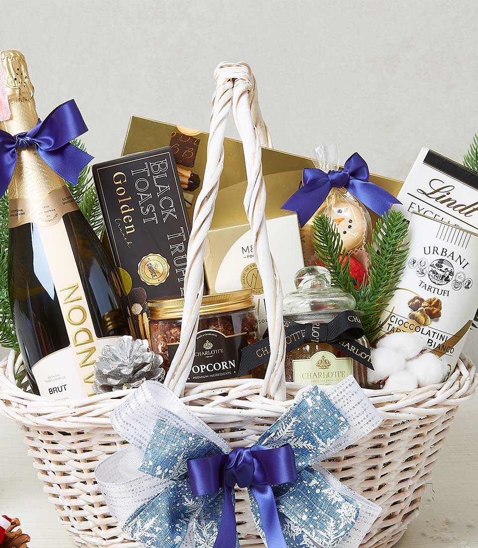 A luxurious white wicker gift basket, meticulously curated by Basketeer, brimming with an assortment of gourmet delights. Nestled within are a bottle of exquisite sparkling wine, decadent boxes of chocolates, artisanal popcorn, and a sumptuous Lindt chocolate bar. The basket is elegantly adorned with blue ribbons showcasing the Basketeer logo, accompanied by a festive pine cone and lush evergreen branches, creating an inviting holiday ambiance.