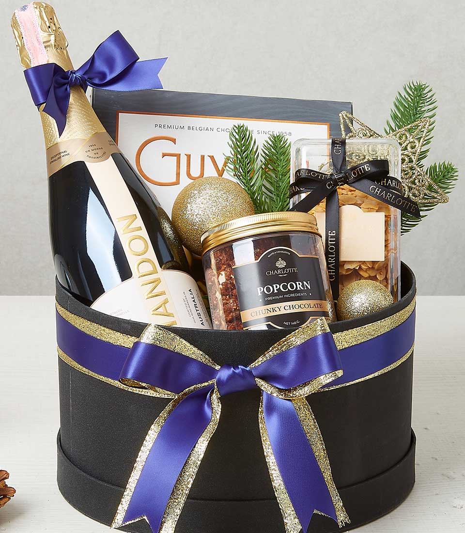 A luxurious holiday gift basket elegantly curated by Basketeer, featuring a bottle of opulent Chandon champagne, an exquisite box of Guylian chocolates, a premium container of chocolate caramel popcorn, and a sparkling star-shaped ornament. The ensemble is complemented by radiant gold baubles, all meticulously arranged in an elegant black box adorned with sophisticated gold trim and tied with a sumptuous purple ribbon bearing the Basketeer logo.