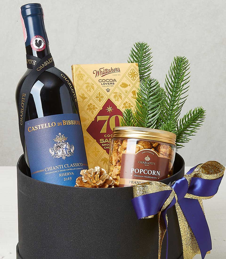 A luxurious Basketeer gift box presenting an exquisite bottle of Castello di Bibbione Chianti Classico wine, complemented by a decadent bar of Whittaker's 70% cocoa dark chocolate and a delightful jar of Charlotte's gourmet popcorn. The scene is elegantly adorned with a charming pine branch and a glistening gold pine cone, while the box itself is beautifully wrapped in opulent blue and gold ribbons, showcasing the Basketeer logo.