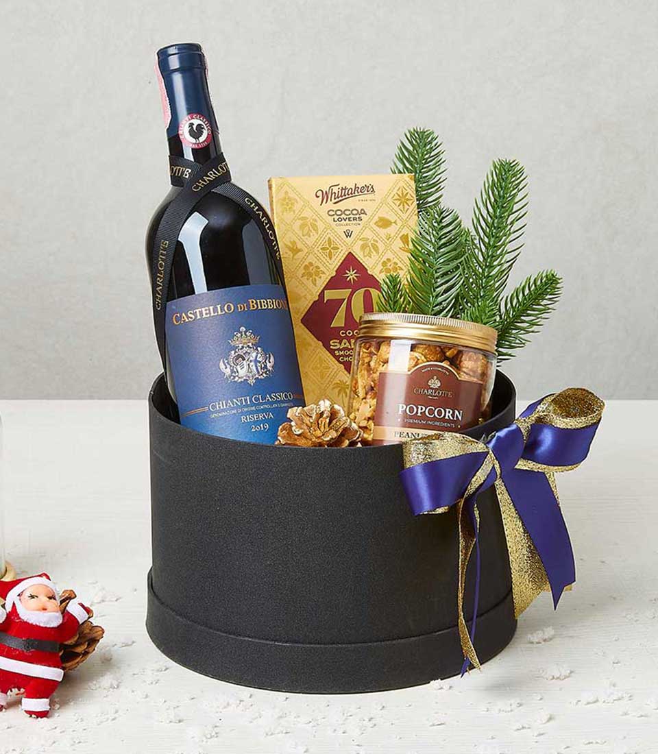 An exquisite gift basket by Basketeer showcasing a bottle of the elegant Castello di Bibbione Chianti Classico wine, a indulgent box of Whittaker's milk chocolate, and a jar of artisanal caramel popcorn. Accentuated with a charming small pine branch and decorative pine cone, all tastefully arranged in an opulent black box adorned with Basketeer's signature purple and gold ribbon.
