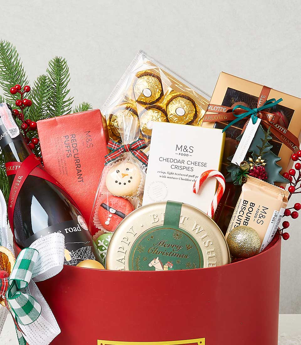 A luxurious red gift basket from Basketeer, elegantly filled with an assortment of premium holiday treats. Relish the indulgence of Ferrero Rocher chocolates, savor the savory cheddar cheese crispies, delight in red currant puffs, and enjoy a bottle of fine wine. Beautifully decorated biscuits and a tin of exquisite cookies add to the festive spirit. The basket is adorned with lush greenery and vibrant berries for an inviting cheerful look. A sophisticated touch is added with Basketeer's signature ribbon and branding seamlessly integrated into the packaging.
