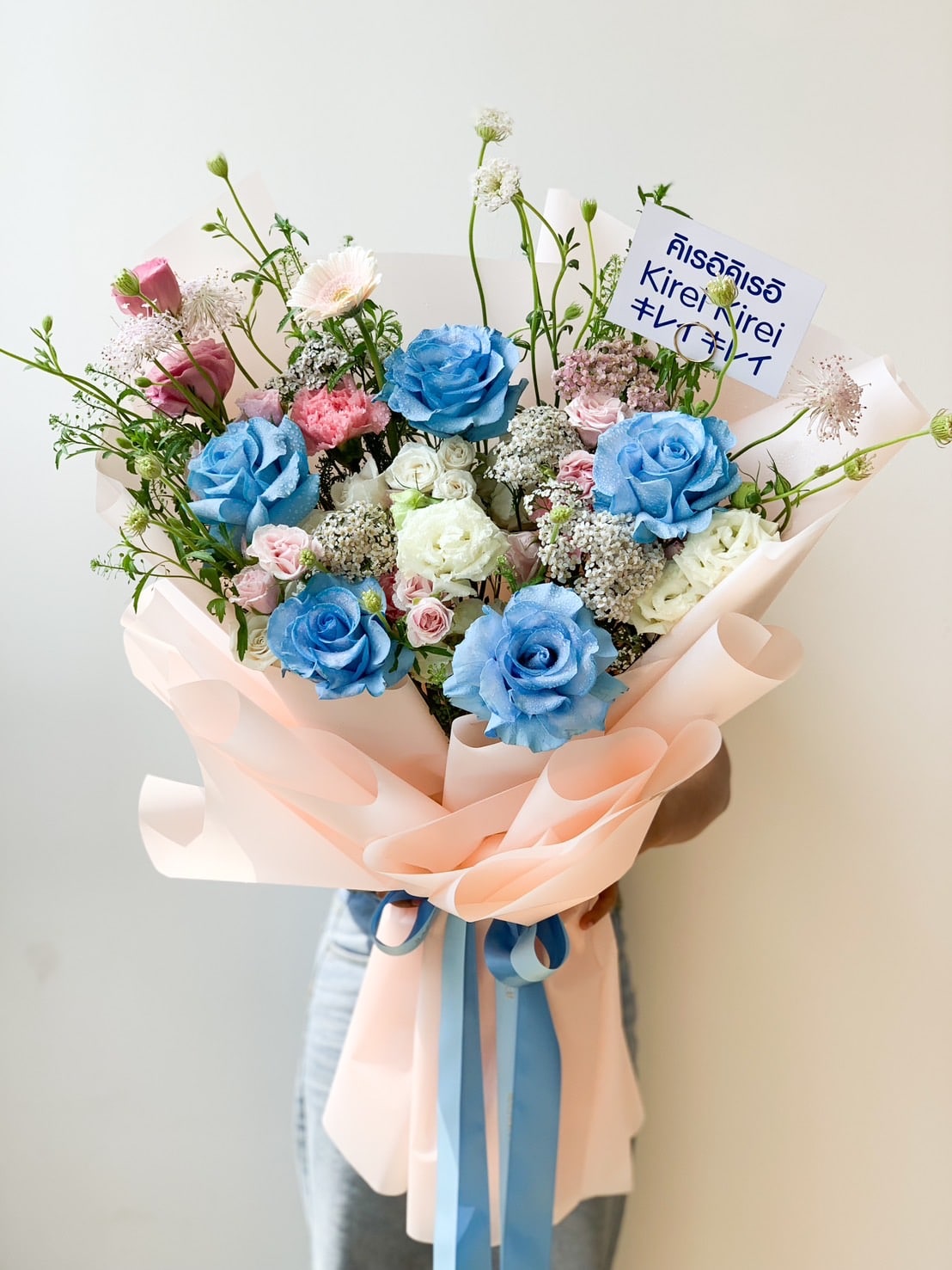 A vibrant blossoms bouquet featuring blue and white roses, baby's breath, and small pink flowers, wrapped in peach-colored paper with matching blue and peach ribbons. The bouquet includes a card with text written in different languages.