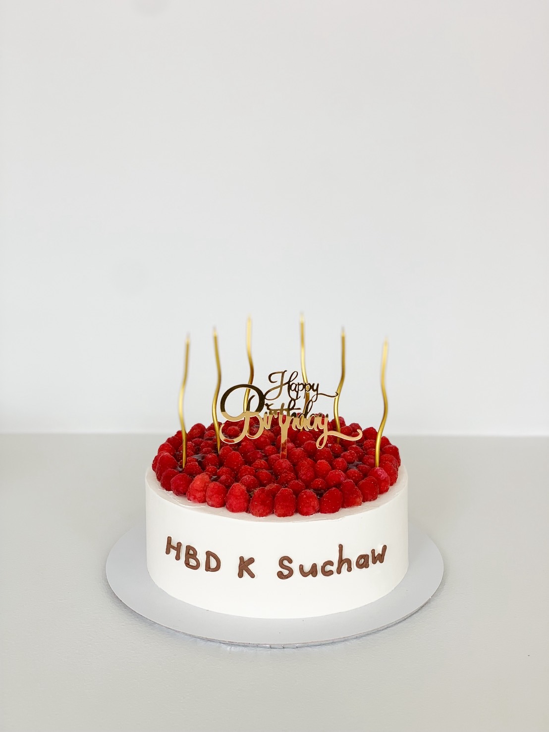 White minimalist birthday cake topped with fresh raspberries, gold swirl candles, and “Happy Birthday” topper