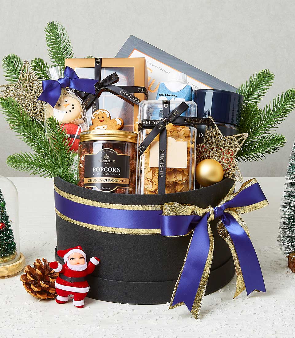 A luxurious festive gift basket curated by Basketeer, brimming with an opulent assortment of delights. Nestled within a sophisticated black round basket draped in an elegant blue ribbon bearing the Basketeer logo, you'll find a jar of gourmet popcorn, a decadent box of artisan chocolates, and an exquisite star-shaped ornament. Completing this enchanting scene are a charming Santa figurine and a beautifully crafted holiday-themed snow globe, creating an inviting and sumptuous display perfect for the season.