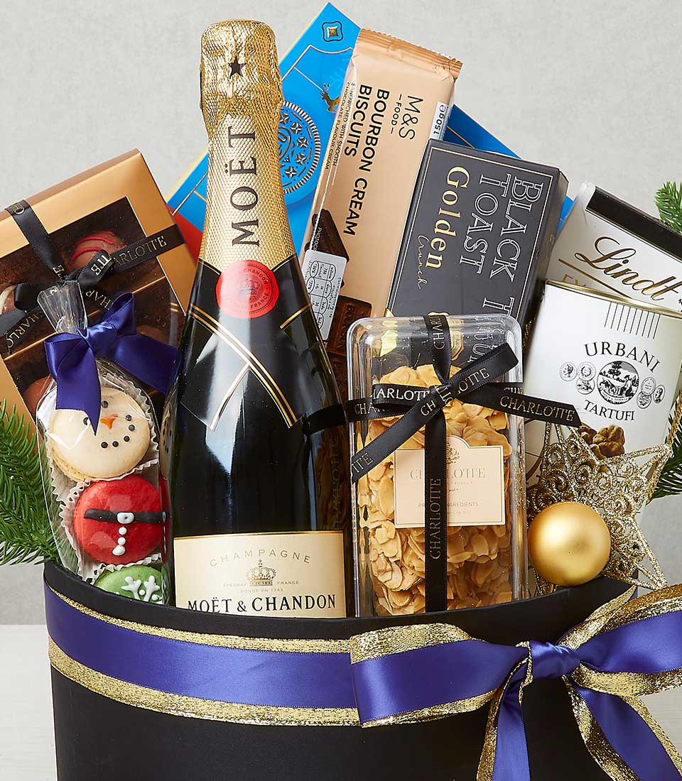 A luxurious gift basket curated by Basketeer, featuring an exquisite bottle of Moët & Chandon champagne, an assortment of decadent Lindt chocolates, and gourmet M&S biscuits. Black Toast crackers add a touch of sophistication to this indulgent collection. The basket is elegantly adorned with a sumptuous purple and gold ribbon, proudly showcasing the Basketeer logo as a mark of quality and taste. Decorative items enhance the festive ambiance, making this gift an inviting treasure for any special occasion.