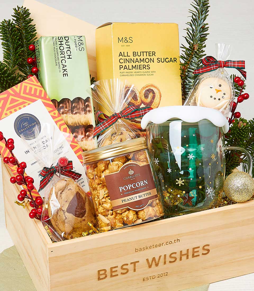 A luxurious Basketeer gift basket teeming with premium delights: M&S Dutch shortcake, buttery cinnamon sugar palmiers from M&S, an elegantly iced cookie, delectable peanut butter popcorn, a charming green mug adorned with a festive holiday motif, a shimmering gold ornament, and beautifully arranged sprigs of evergreen with vibrant red berries. All these exquisite items are meticulously presented with the Basketeer logo embellishing the ribbon and box for that added touch of opulence.