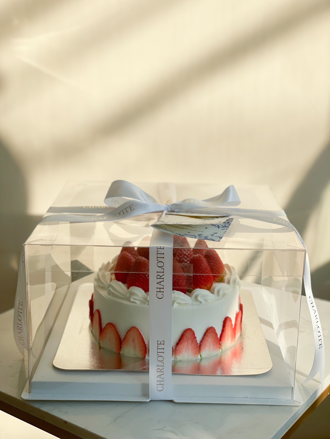 A beautifully decorated strawberry shortcake with fresh strawberries and whipped cream, elegantly packaged in a transparent gift box with a white ribbon.