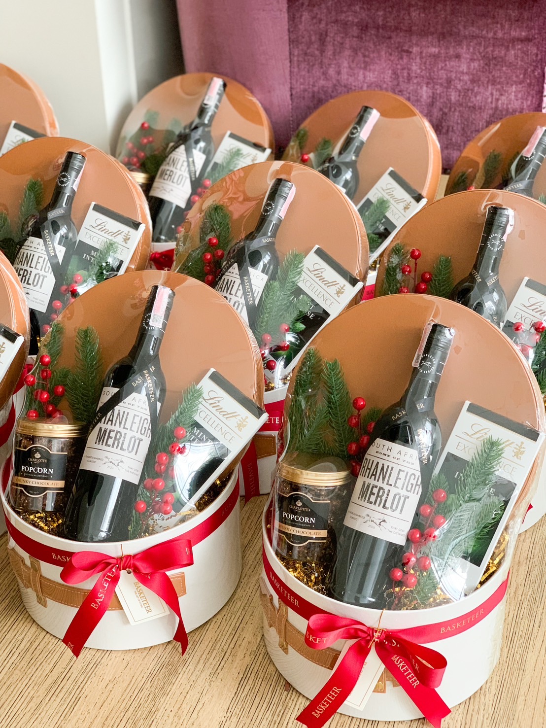 A lavish gift basket from Basketeer featuring a fine bottle of red wine, an exquisite jar of gourmet popcorn, and a decadent Lindt chocolate bar. The basket is elegantly adorned with lush evergreen sprigs and vibrant red berries. A delicate ribbon showcases the Basketeer logo, adding an extra touch of sophistication. The scene is enriched by a charming Santa figurine and festive holiday decorations in the background, making it irresistibly inviting.