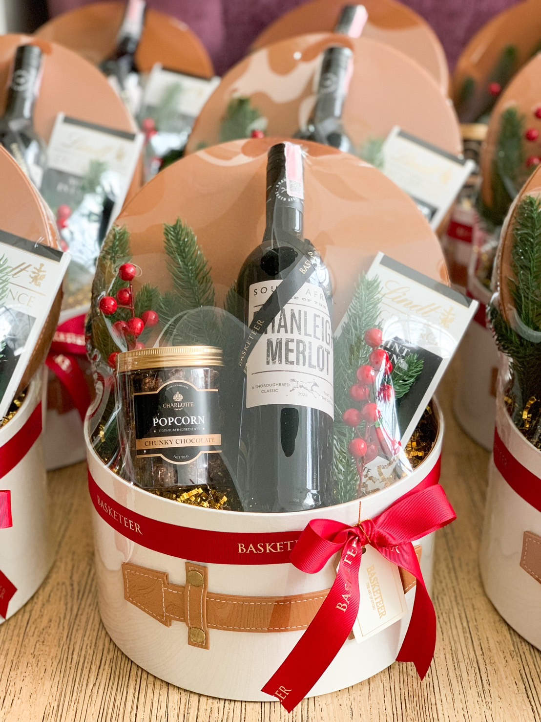 A lavish gift basket from Basketeer featuring a fine bottle of red wine, an exquisite jar of gourmet popcorn, and a decadent Lindt chocolate bar. The basket is elegantly adorned with lush evergreen sprigs and vibrant red berries. A delicate ribbon showcases the Basketeer logo, adding an extra touch of sophistication. The scene is enriched by a charming Santa figurine and festive holiday decorations in the background, making it irresistibly inviting.