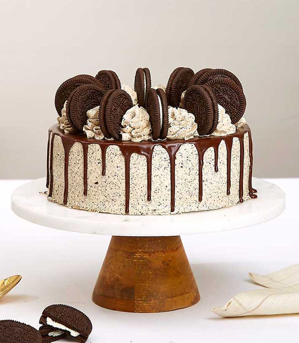 An exquisite Oreo-Chocolate Cream Cake from Basketeer rests elegantly on a polished wooden cake stand. The decadent creation is crowned with whole chocolate sandwich cookies and adorned with sumptuous dollops of whipped cream. A luxurious chocolate drizzle cascades gracefully around the edges, while additional cookies temptingly flank the scene, inviting indulgence in this lavish confectionery delight. The signature Basketeer ribbon and box add an extra touch of sophistication to this opulent presentation.