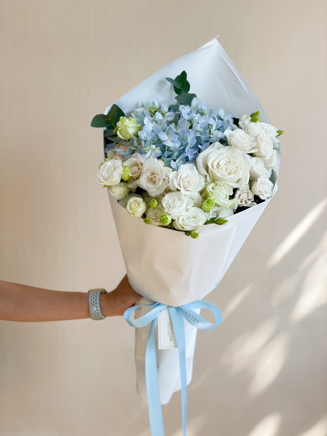 A beautifully arranged white and blue bouquet featuring white roses, lisianthus, and blue hydrangeas, wrapped in white paper with a blue ribbon.