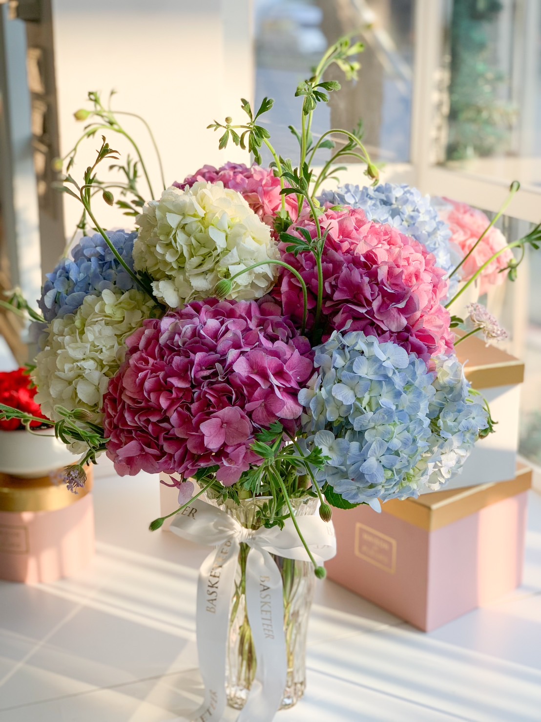 A gorgeous pastel hydrangea bouquet arranged in a clear glass vase, tied with a luxurious Basketeer ribbon, perfect for gifting or home decoration.