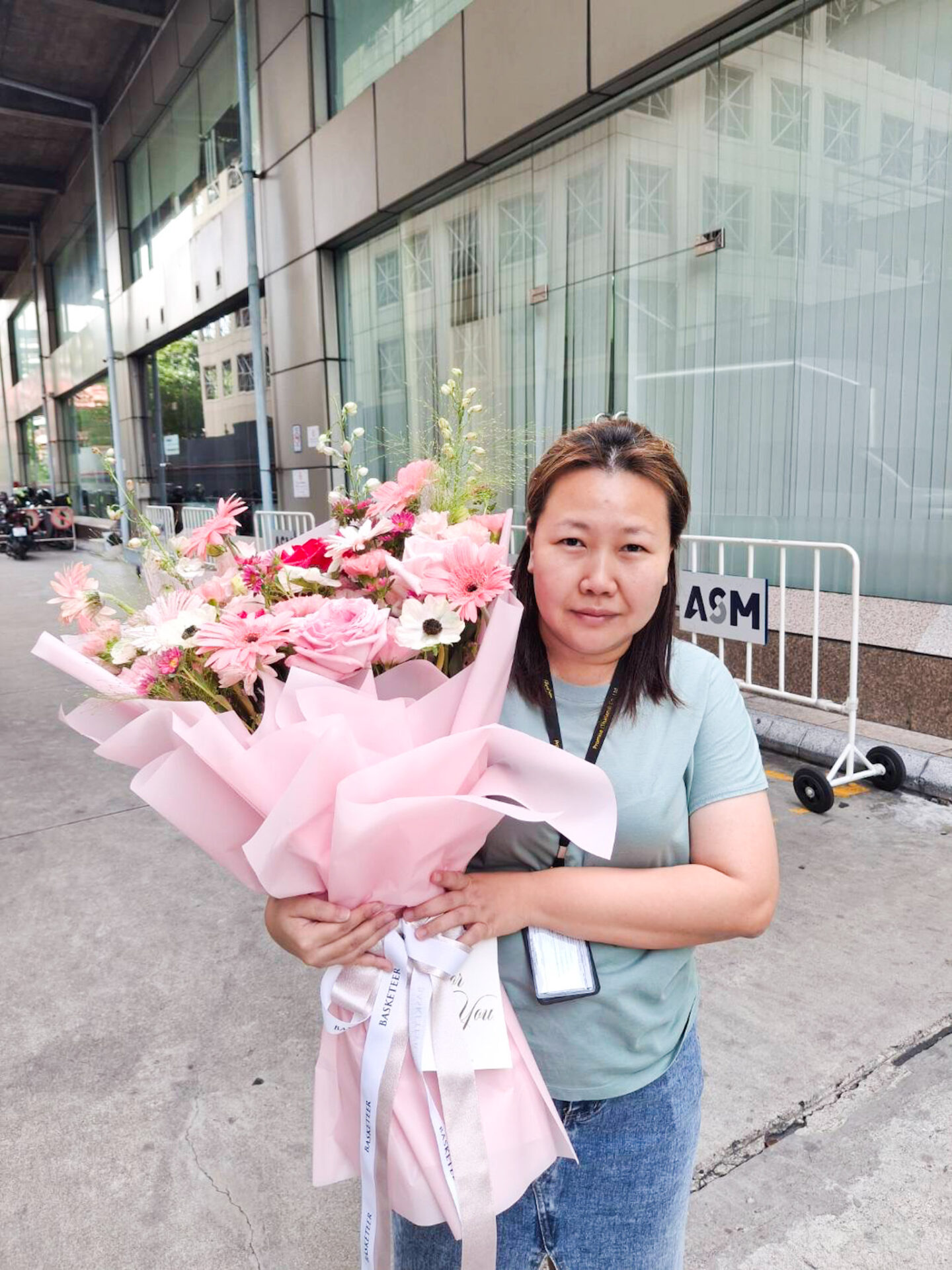 A woman, elegantly dressed in a light green blouse paired with chic jeans, radiates grace as she stands outdoors cradling an opulent bouquet of delicate pink and white flowers, exquisitely wrapped in luxurious pink paper. The sophisticated ambiance is complemented by the backdrop of a sleek modern building adorned with gleaming glass panels. Nearby, under a gracefully covered walkway, hints of the prestigious Basketeer brand can be seen on ribbon accents and gift boxes, adding a touch of refined elegance to the scene.