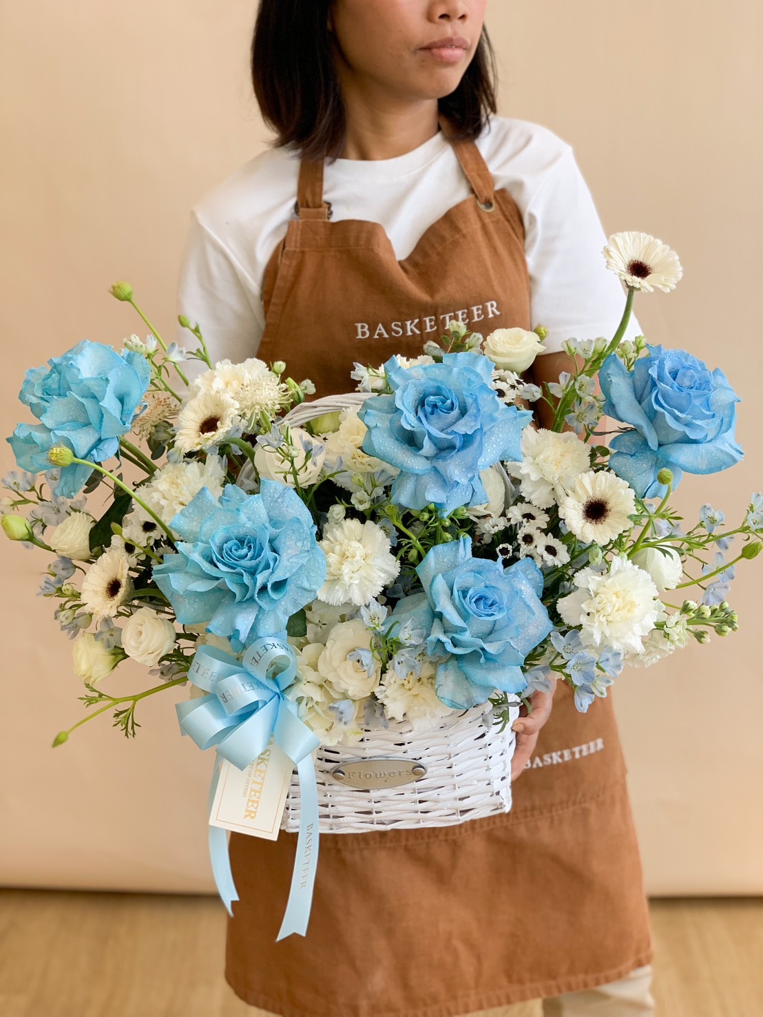 A stunning floral arrangement featuring blue roses, white blooms, and lush greenery, elegantly placed in a white wicker basket with a soft blue ribbon. A timeless gift for birthdays, anniversaries, baby showers, and special celebrations.