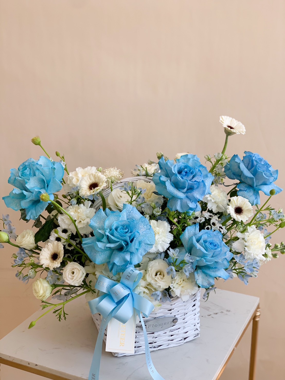 A luxurious blue and white floral arrangement featuring blue roses, white blooms, and soft greenery elegantly placed in a white wicker basket with a delicate blue ribbon. A sophisticated gift for birthdays, anniversaries, baby showers, and special occasions.