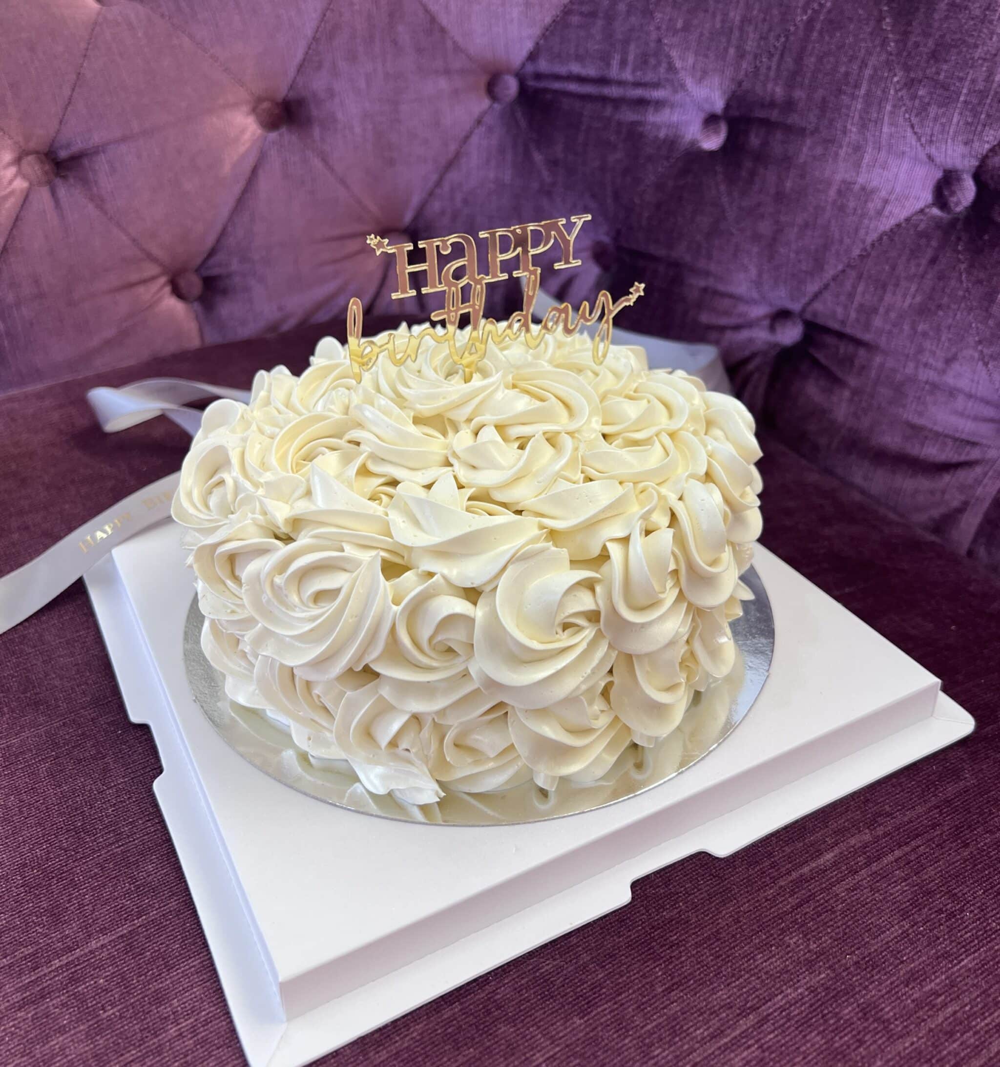 A round white cake, aptly named the Creamy Indulgence Cake, decorated with intricate cream swirls and topped with a gold 