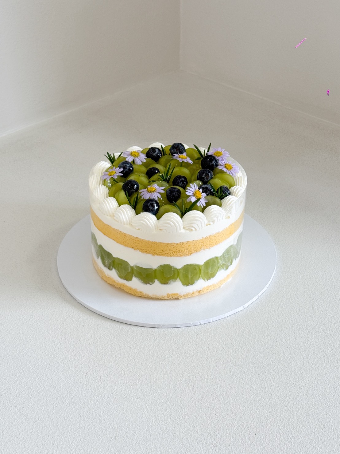 A round Grape & Blueberry Birthday Cake decorated with white icing, green grapes, blueberries, and small white flowers on top, placed on a white cake board against a plain background.