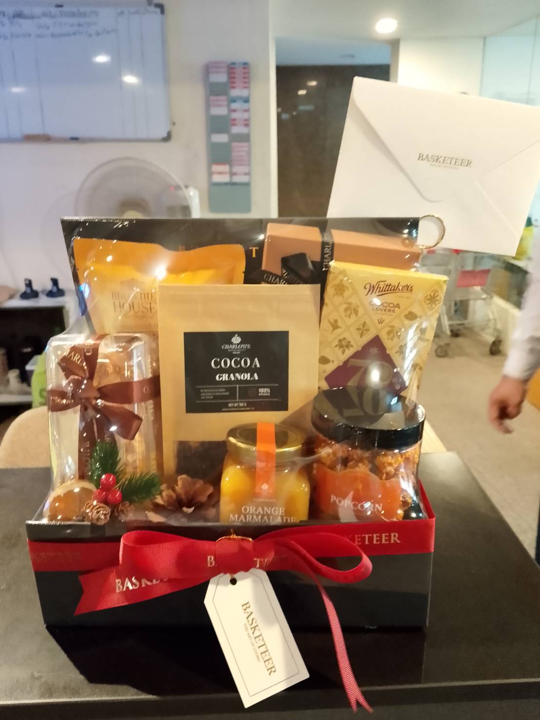 The Epicure’s Delight Gift Ensemble features cocoa granola, orange marmalade, popcorn, chocolate, a gold bow, red ribbon, and a white envelope, all beautifully displayed indoors on a worktop.