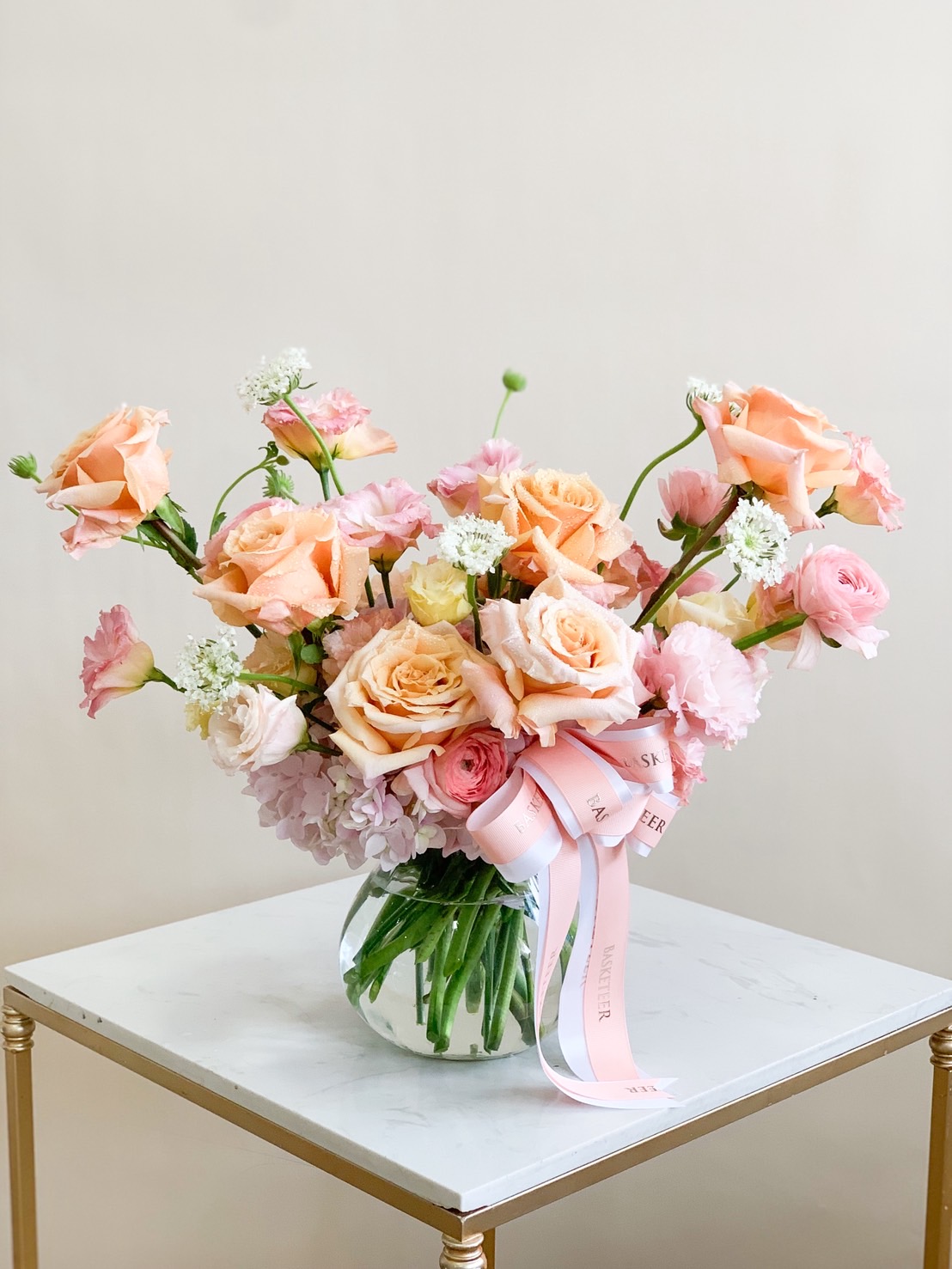 Flower arrangement in a glass vase with peach roses, pink lisianthus, ranunculus, and white filler flowers tied with pastel pink ribbons.
