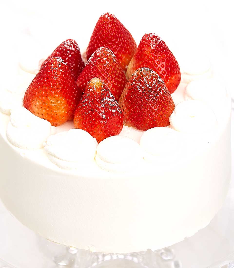 A luxurious round white cake adorned with plump, succulent strawberries and artistically piped dollops of velvety whipped cream along the edges. The creamy masterpiece is presented against a pristine white backdrop, accentuating its sophisticated elegance. A ribbon bearing the 