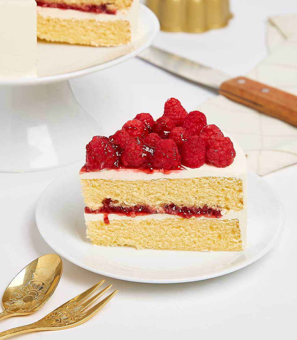 A sumptuous slice of Basketeer’s Temptation Cake graces a pristine white plate, adorned with succulent raspberries and lavish raspberry jam. Its creamy frosting and decadent layer of raspberry filling beckon irresistibly. Nearby, an exquisite gold fork and spoon set await indulgence, while the remaining masterpiece stands proudly on a luxurious cake stand in the background, enveloped by the elegance of Basketeer's signature style.