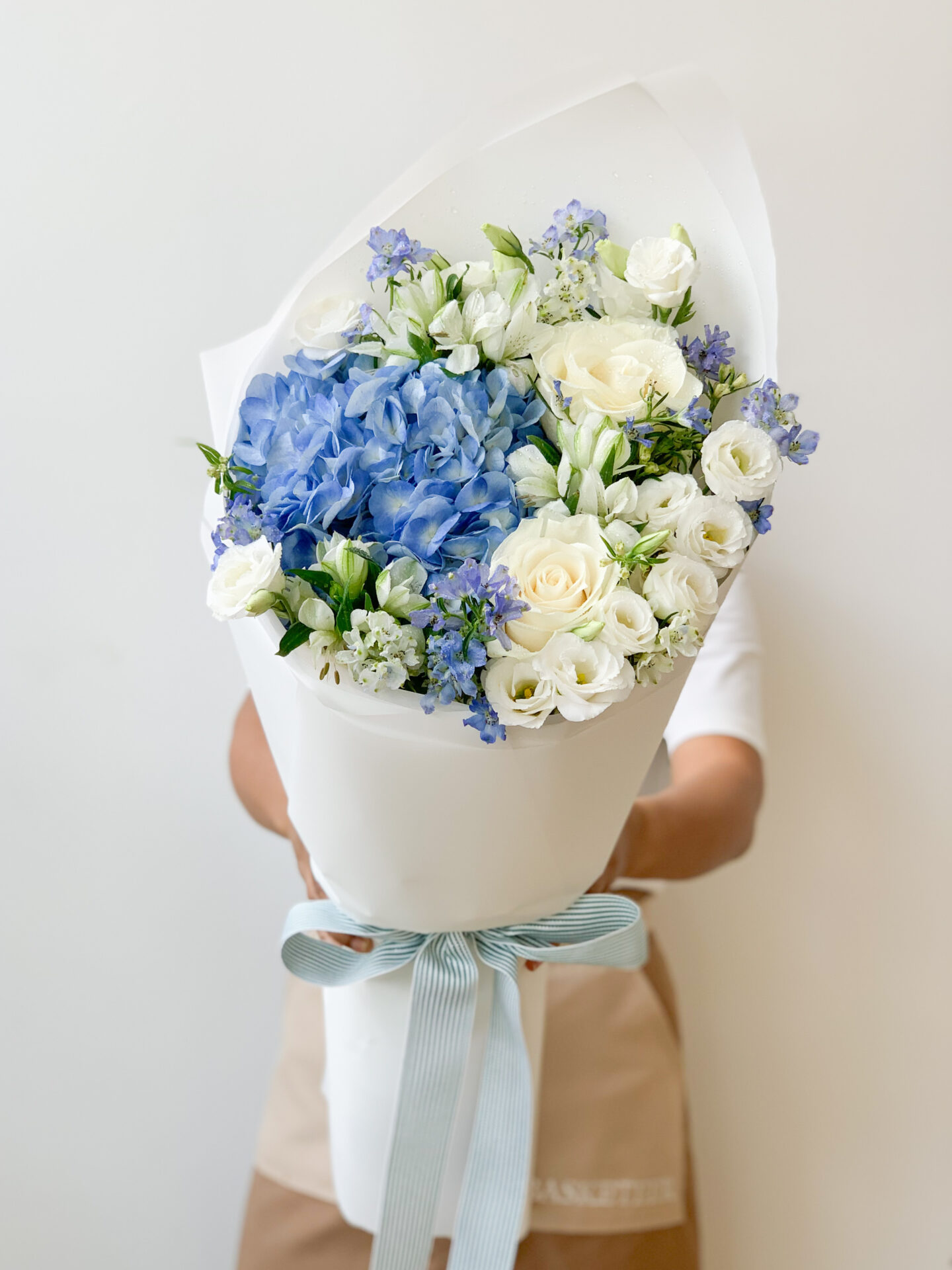 A person holds a large bouquet of flowers wrapped in white paper. The bouquet includes blue and white flowers, predominantly roses and hydrangeas, with green foliage. The bouquet is tied with a light blue ribbon. The background is plain white.