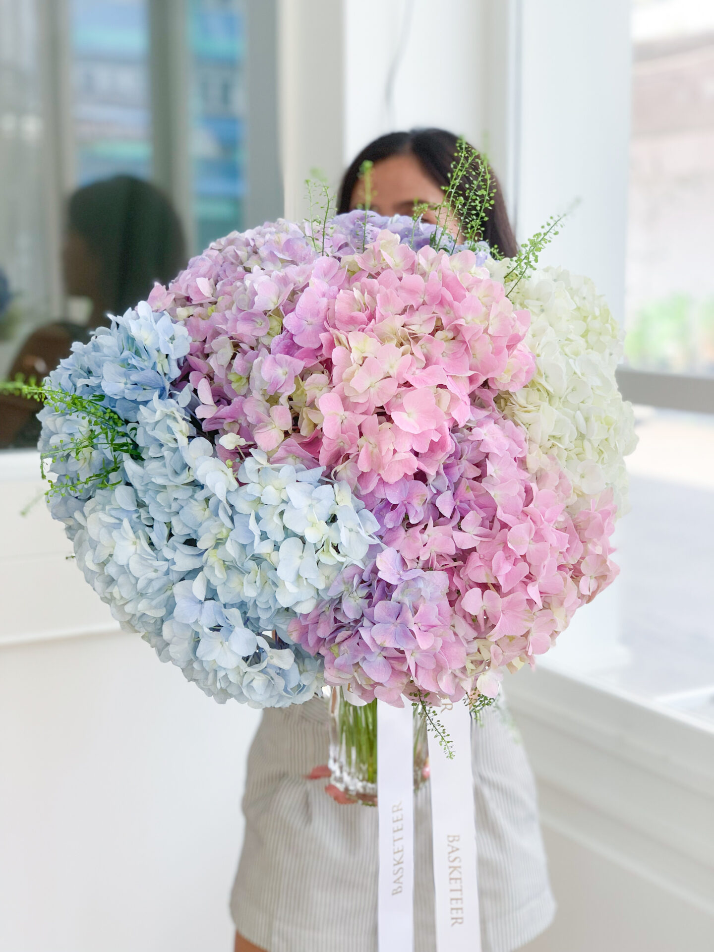 A person stands behind a large, elegant vase filled with hydrangeas in varying shades of blue, pink, and white. The big bouquet obscures most of their face and upper body. The person is indoors, wearing striped clothing, and light filters through a window in the background.