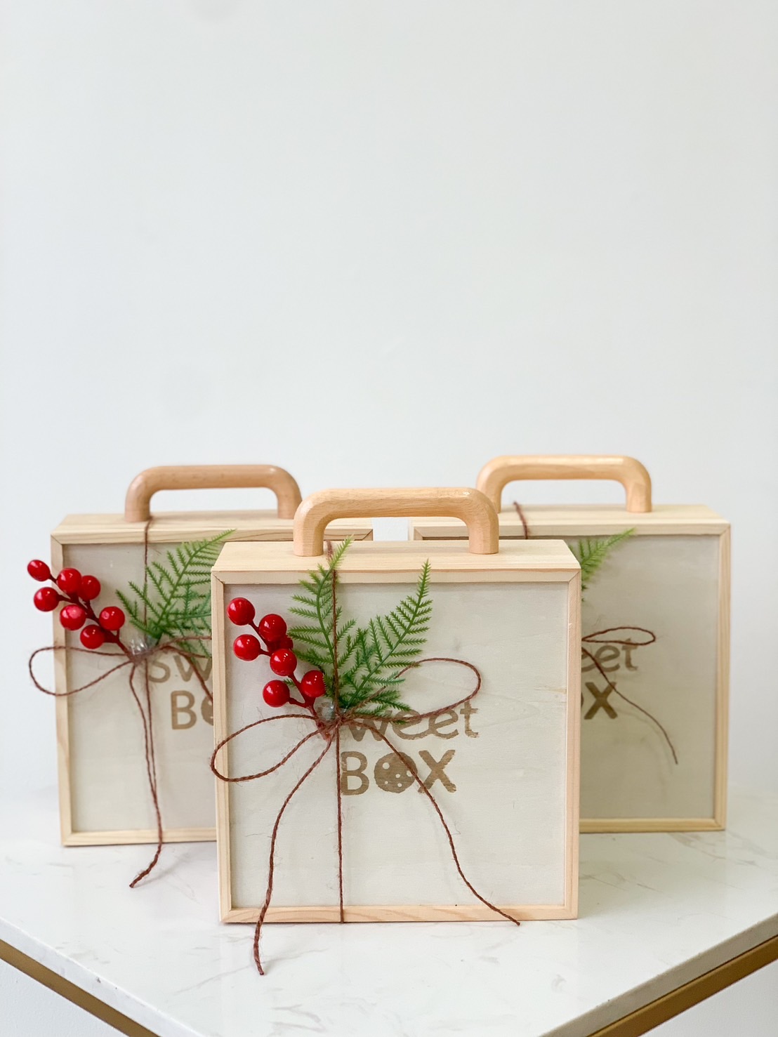 Three wooden gift boxes with handles, adorned with red berries and green fern fronds tied with twine, are arranged on a white surface. The text 