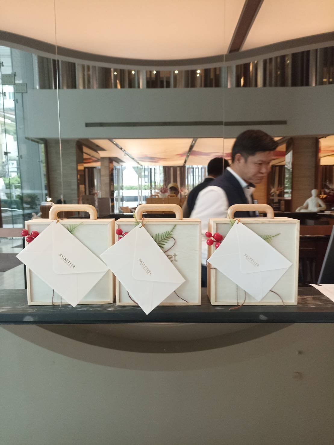 Three neatly arranged boxed gifts with envelopes on top, adorned with decorative greenery and red berries, are displayed on a counter. A server in a black vest and white shirt walks in the background under a modern, well-lit interior with wooden accents.