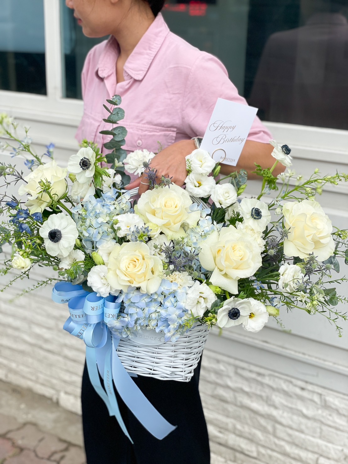 A person in a pink shirt holds Love’s Blue Flowers Gift Basket filled with a beautifully arranged bouquet of white and light blue flowers, including roses and hydrangeas. The basket is adorned with a blue ribbon and a card that reads 