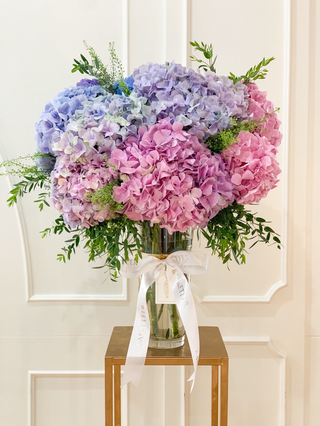 Elegant vase of pastel hydrangeas in pink, purple, and blue shades with fresh greenery, tied with a white ribbon.