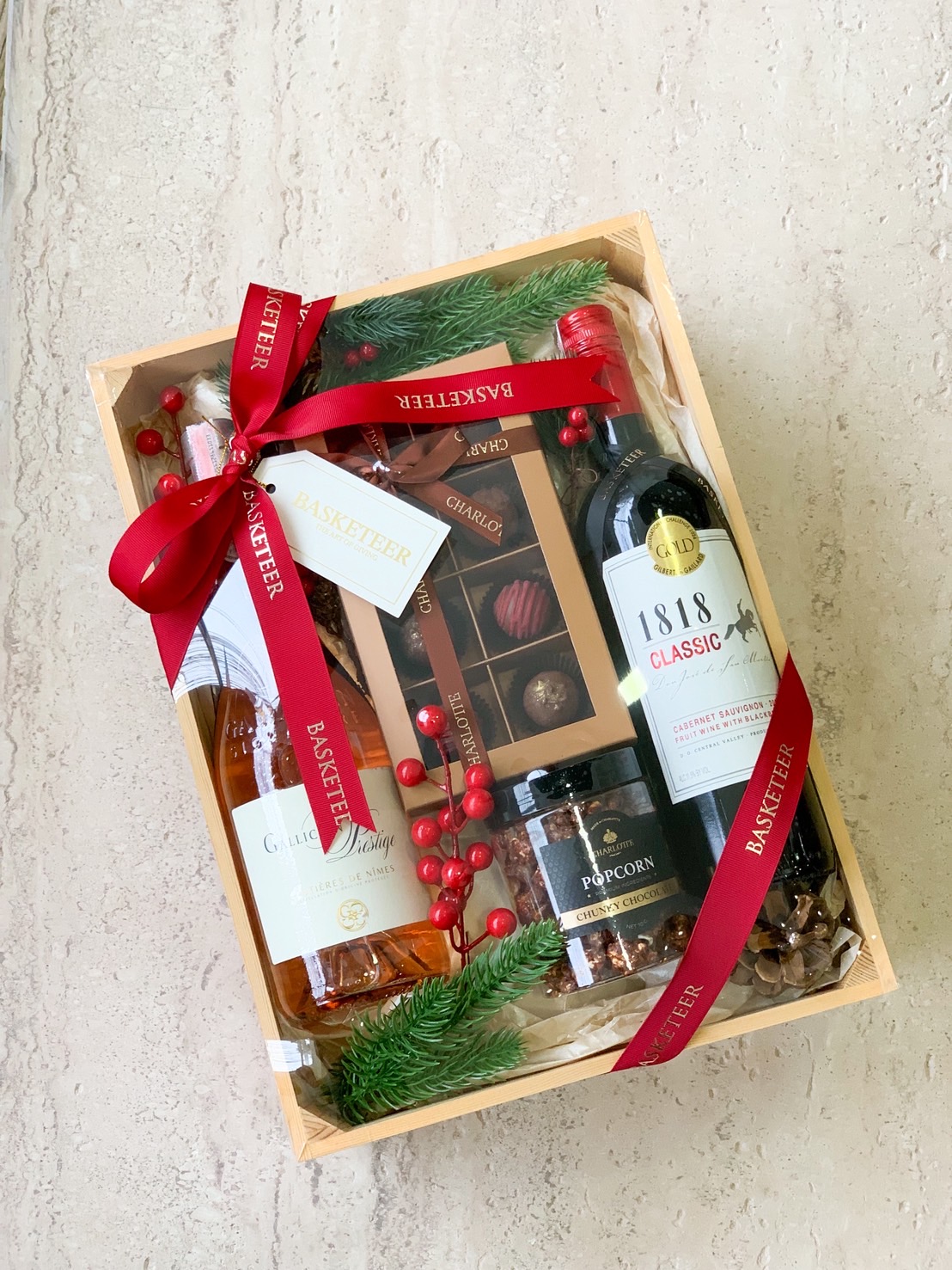 Luxury gift box with red wine, rosé wine, artisanal chocolate, popcorn, and Christmas decor in wooden box with red ribbon.