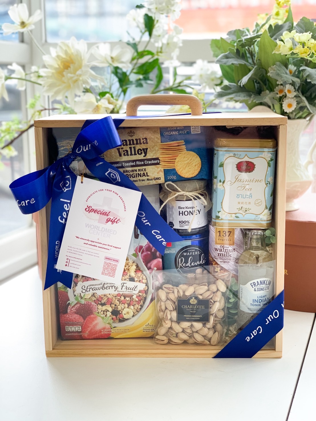 Wooden box filled with health-conscious snacks, herbal drinks, tea, pistachios, and granola, decorated with a navy ribbon and care tag.