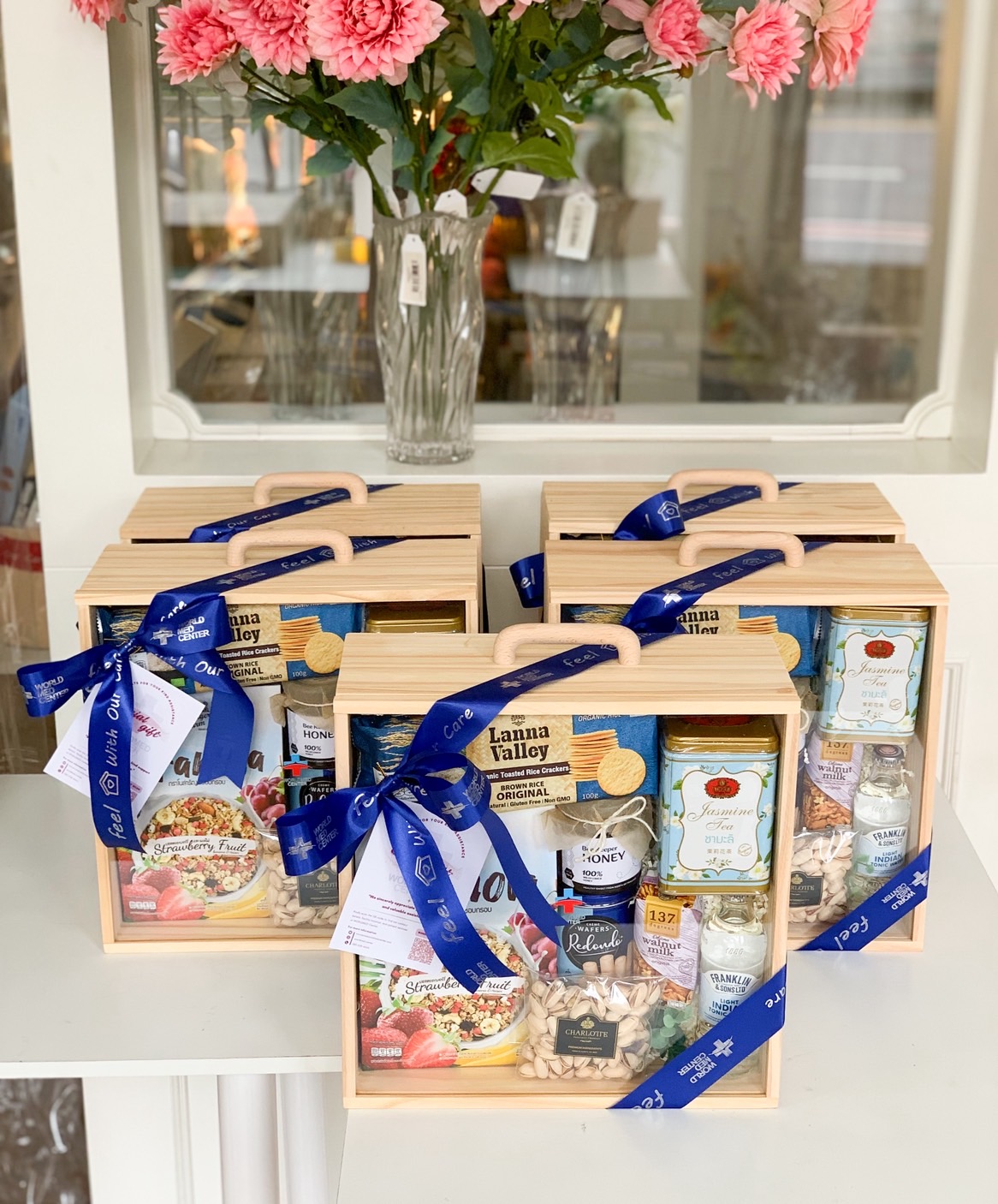 Set of four wooden wellness gift boxes filled with tea, granola, pistachios, honey, and rice crackers, tied with blue corporate ribbon.