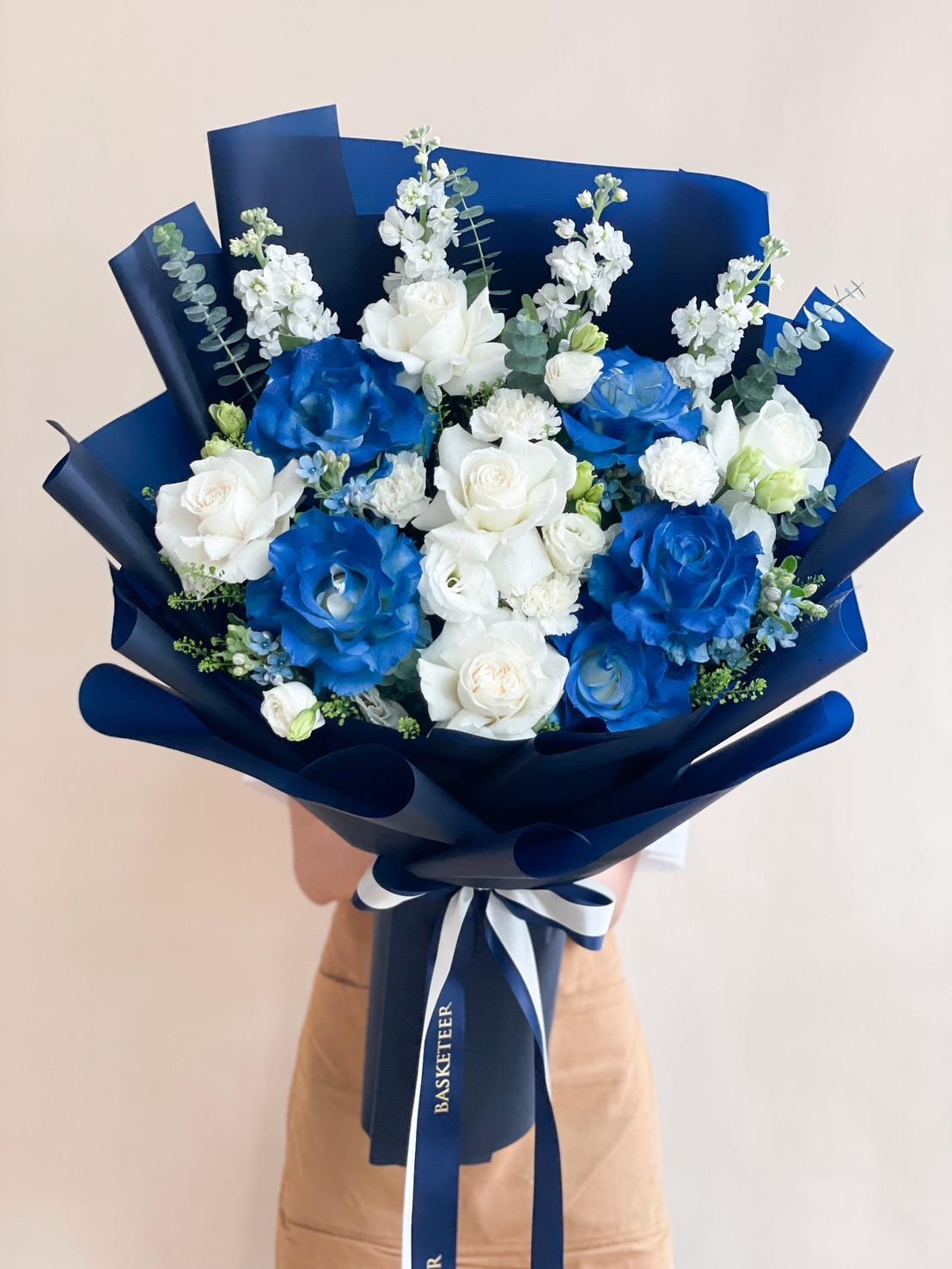 A person gently holds a romantic bouquet consisting of blue and white flowers, accented with green foliage. The blooms are wrapped in dark blue paper and tied with a white ribbon. The bouquet features roses and small flowers, creating an elegant and vibrant arrangement.