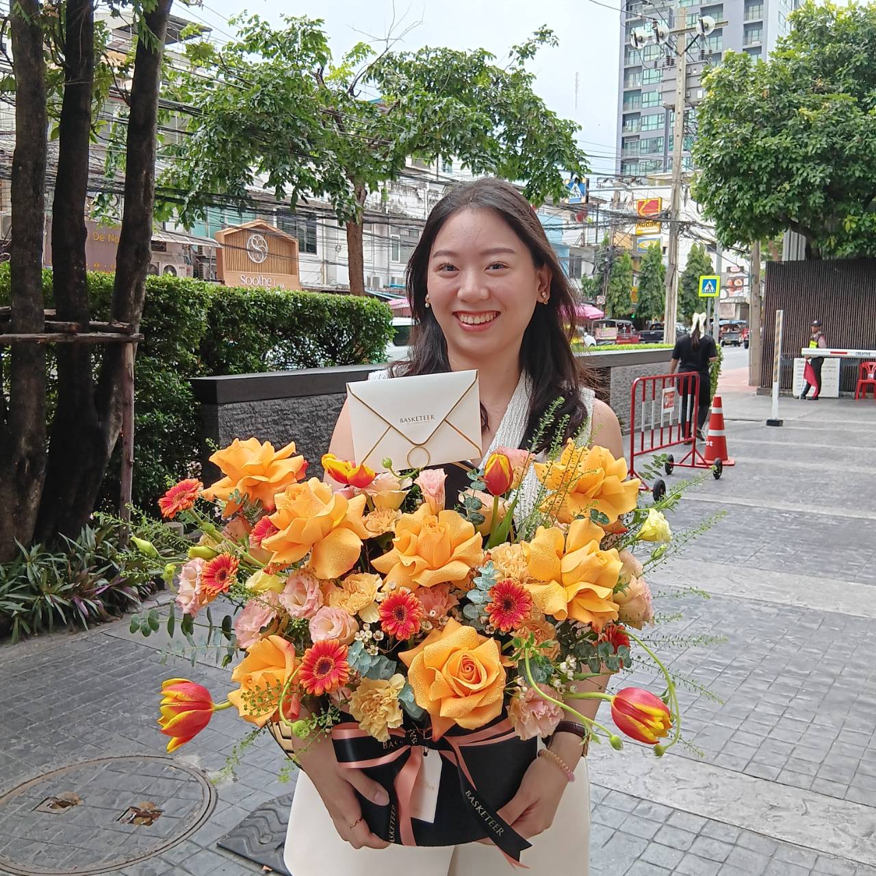 A woman stands on a city sidewalk, smiling while holding a large bouquet of yellow and orange flowers. The background features buildings, trees, and street barriers. She wears a white top and her hair is down.