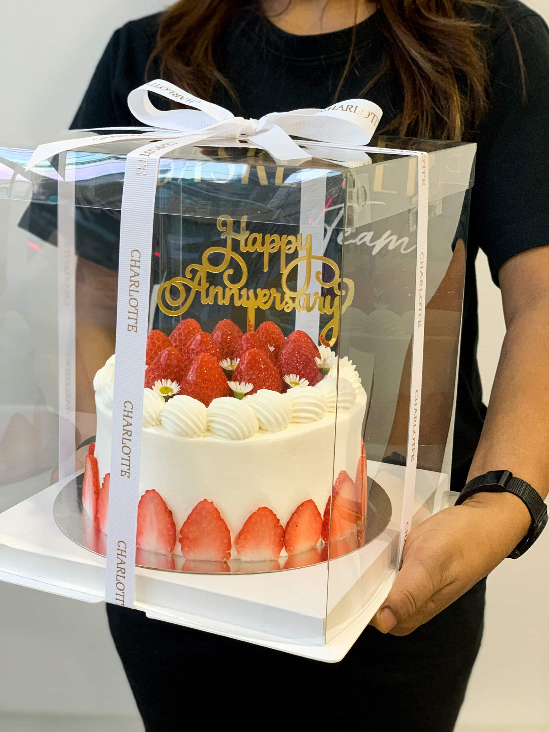 A person is holding a boxed Cream Cake decorated with fresh strawberries and white frosting rosettes. A gold 
