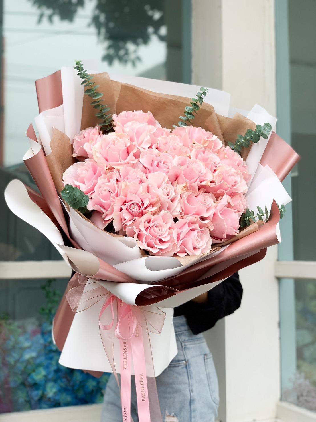 A person holds a large, elegant Pink Mondial Roses Bouquet wrapped in multiple layers of pink and white paper. The bouquet also includes sprigs of greenery, adding a touch of contrast. The background appears to be a window with some green and blue elements visible through it.