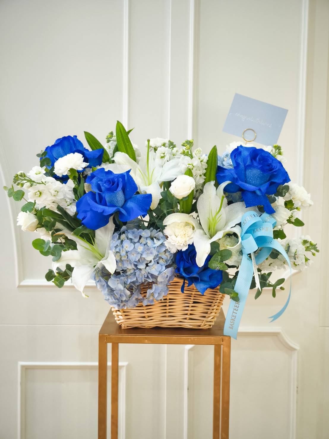 Flower basket with vivid blue roses, white lilies, hydrangeas, and stock flowers, decorated with blue ribbons