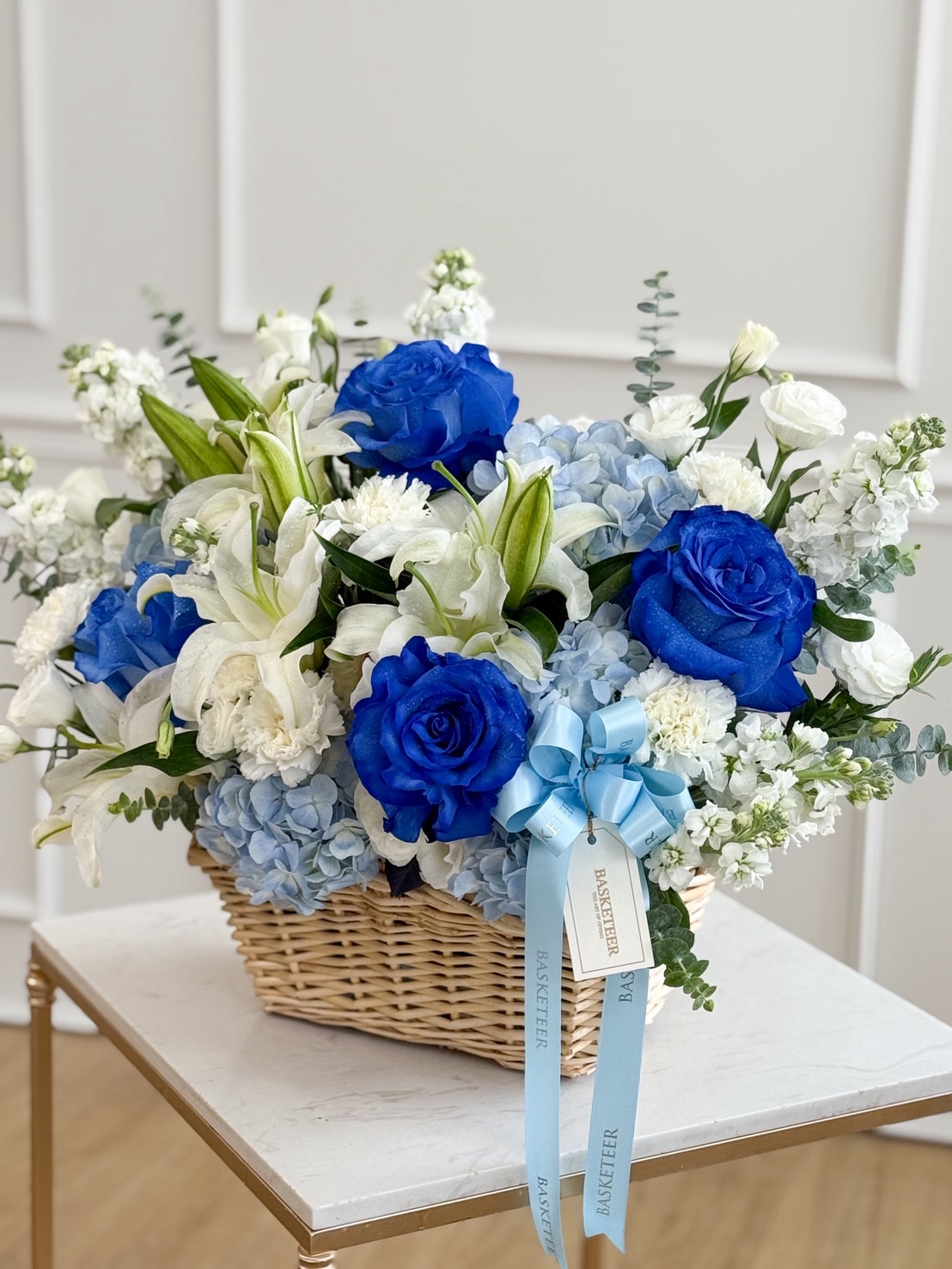The Blue Blooms Celebration Basket features a wicker basket brimming with vibrant blue roses, white lilies, hydrangeas, and assorted white flowers, finished with a blue ribbon and tags, elegantly displayed on a small table against a white wall.