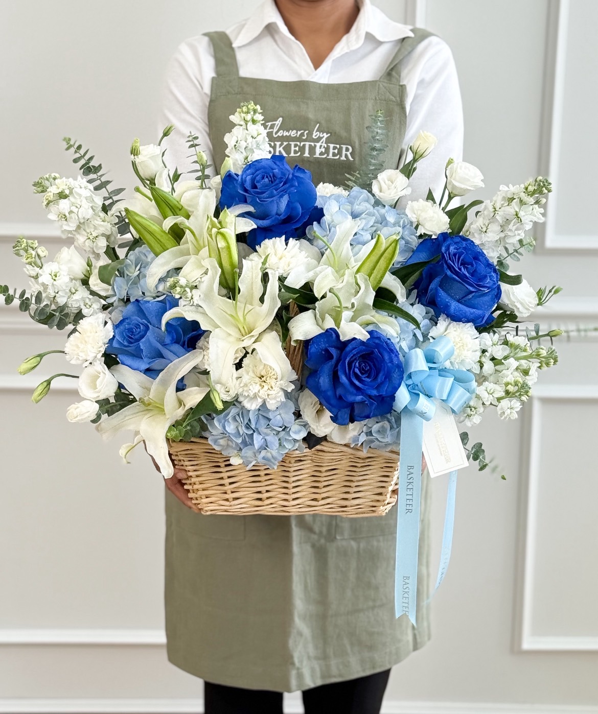 A person in a green apron holds the Blue Blooms Celebration Basket, a wicker basket filled with blue and white roses, lilies, hydrangeas, other white flowers, accented with greenery and a blue ribbon.