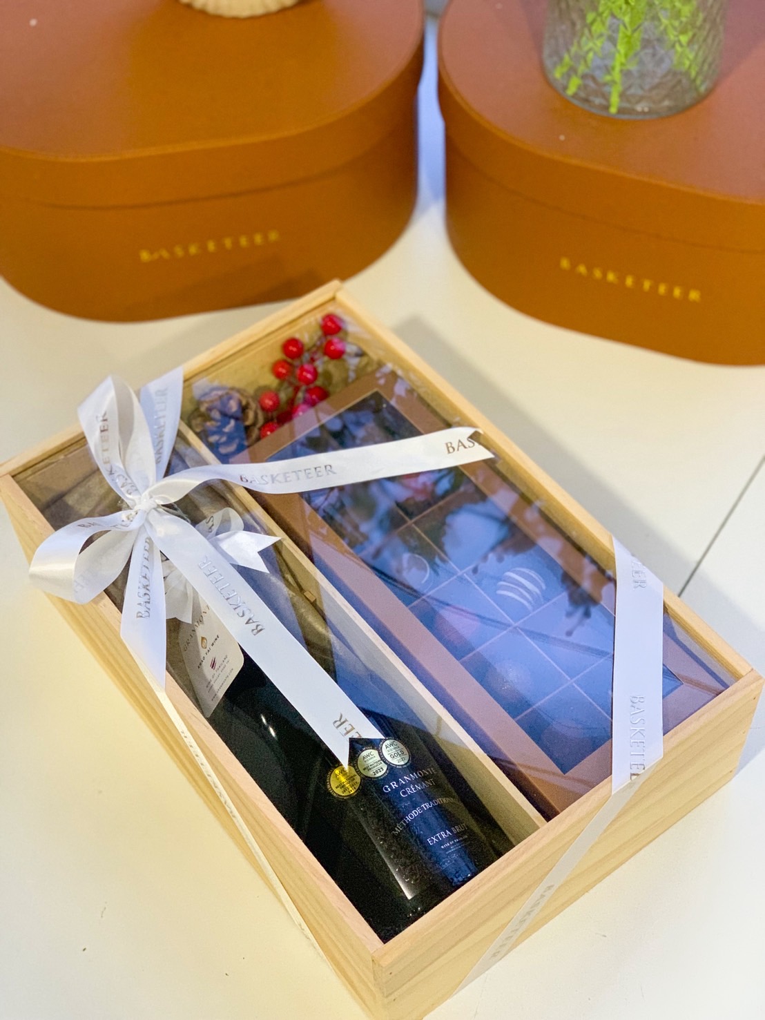 A wooden gift box with a premium bottle of sparkling wine and a box of assorted chocolates tied with a white ribbon.
