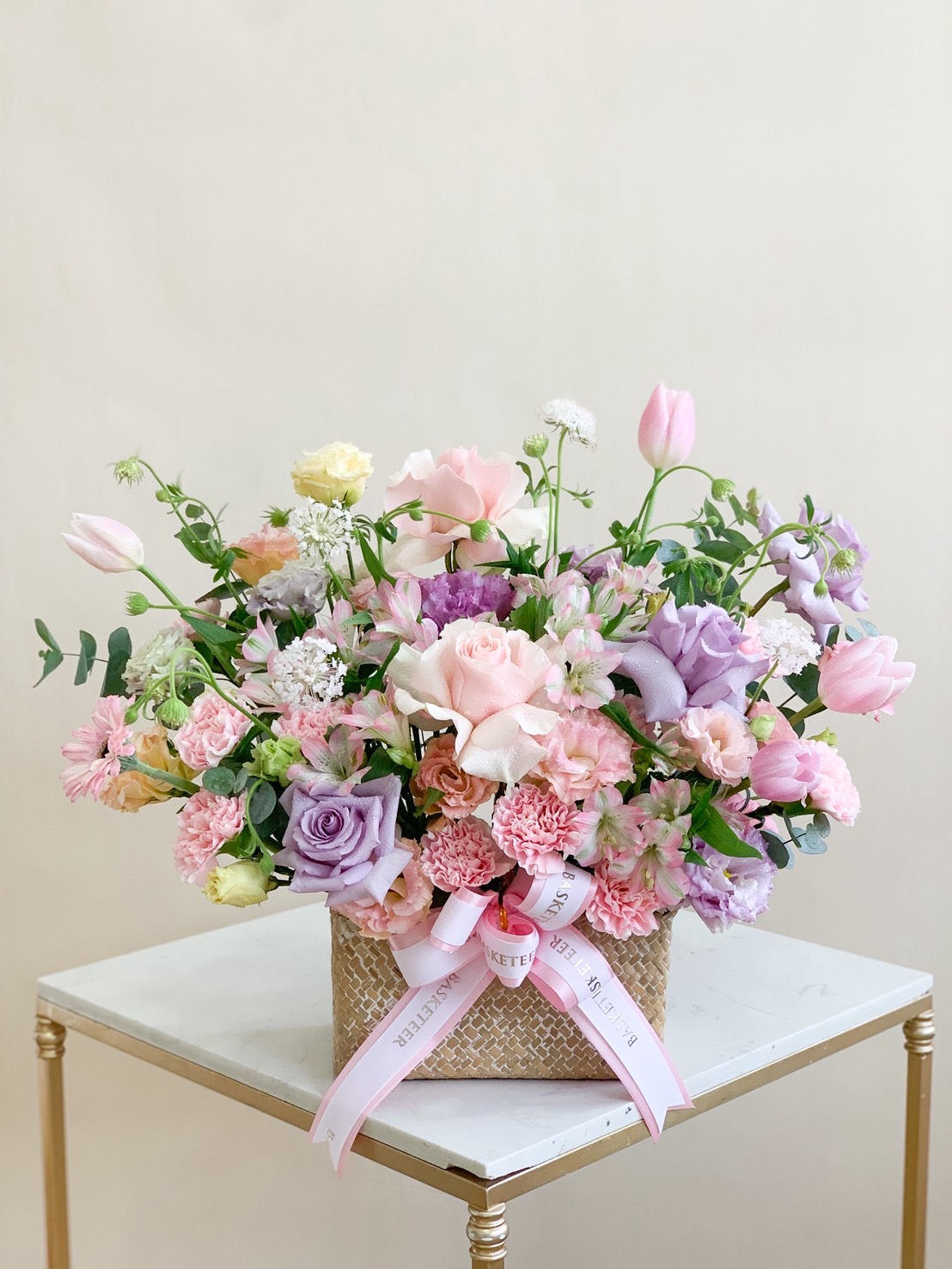 Elegant floral arrangement in a woven box featuring pastel pink, lavender, and peach flowers with lush greenery.