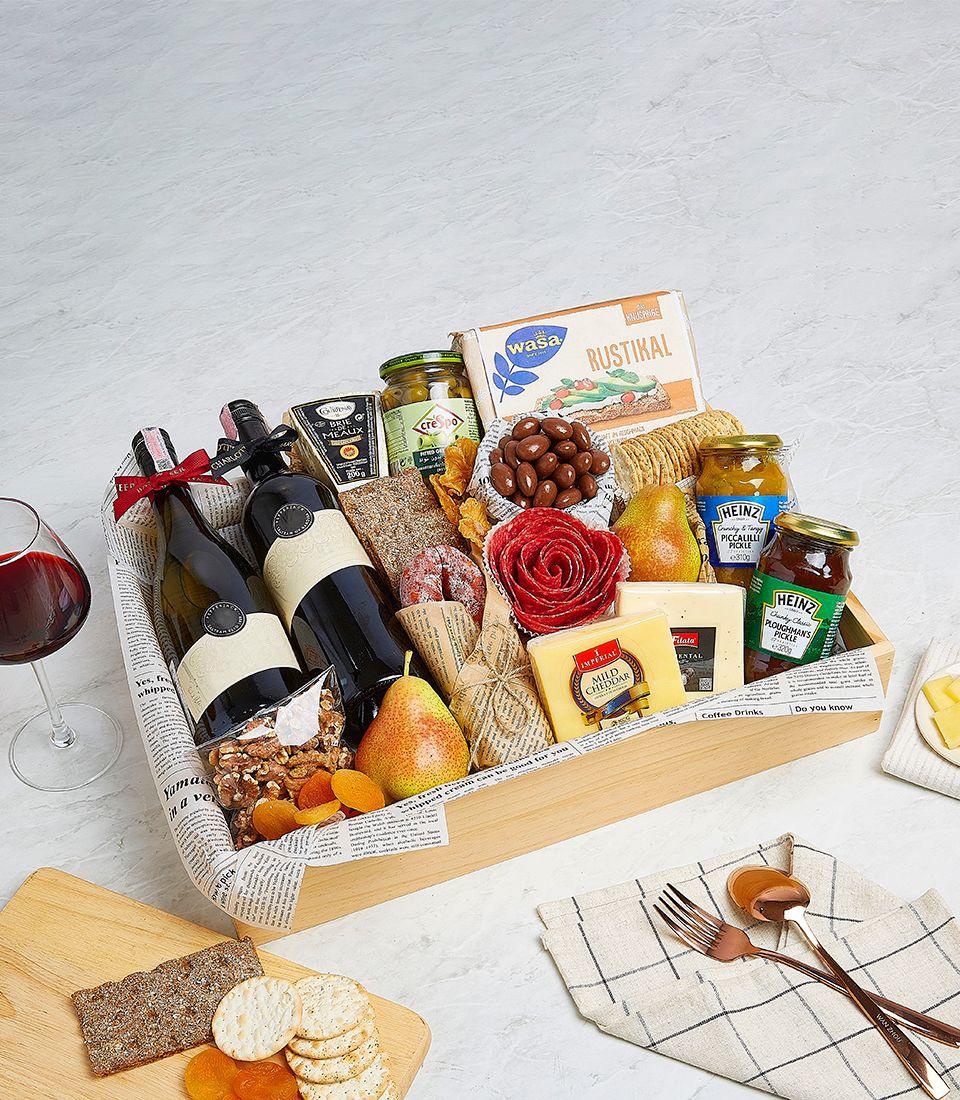 A gift basket contains two wine bottles, assorted cheeses, a red rose-shaped preserved flower, crackers, chocolates, pears, jam, honey, and condiments. A glass of red wine and wooden board with more gourmet snacks are placed beside the basket for perfect wine pairing.
